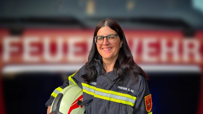 Feuerwehrfrau Eva-Maria Frieser steht in ihrer Einsatzuniform vor einem Feuerwehrauto und hält ihren Helm in der Hand.