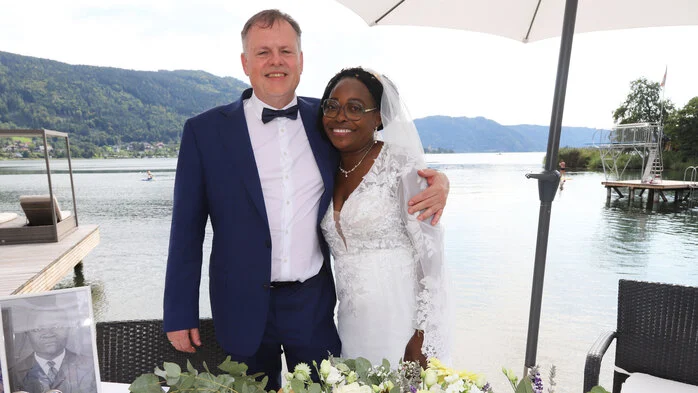 Ein Ehepaar lächelt bei seiner Hochzeit in die Kamera. Im Hintergrund sieht man den Ossiacher See.