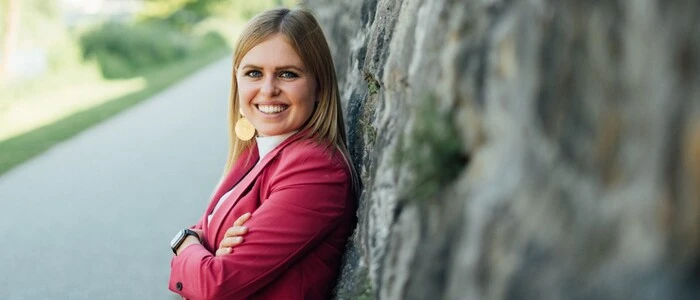 Eine junge Frau, Mitte 20, lehnt sich an eine Steinmauer und lächelt in die Kamera.