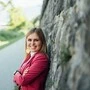 Eine junge Frau, Mitte 20, lehnt sich an eine Steinmauer und lächelt in die Kamera.