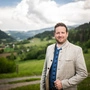 Ein Mann in einem trachtigen Outfit steht vor einer grünen Landschaft und lächelt in die Kamera.