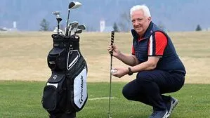 Ein Mann steht auf einem Golfplatz, er hält einen Schläger in der Hand. Daneben steht eine Golftasche mit mehreren Schlägern.