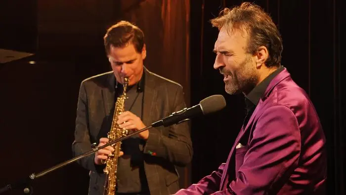 Zwei Musiker, einer am Piano, einer mit Saxophon, stehen auf einer Bühne.