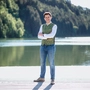 Ein junger Mann, Mitte 20, steht vor einem See und posiert für ein Foto.