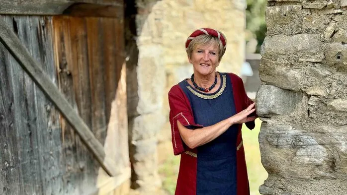 Eine Frau im mittelalterlichen Gewand steht angelehnt an einer Steinmauer.