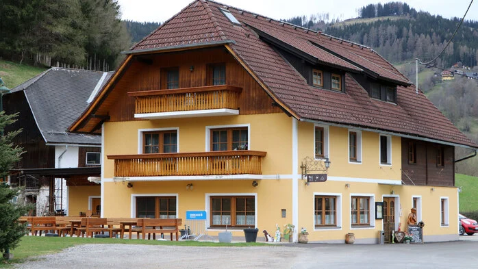 Außenansicht eines großen Bauerngasthauses, eingebettet in Berge und Wälder.