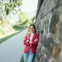 Eine junge Frau, Mitte 20, lehnt an einer Mauer und lächelt in die Kamera.
