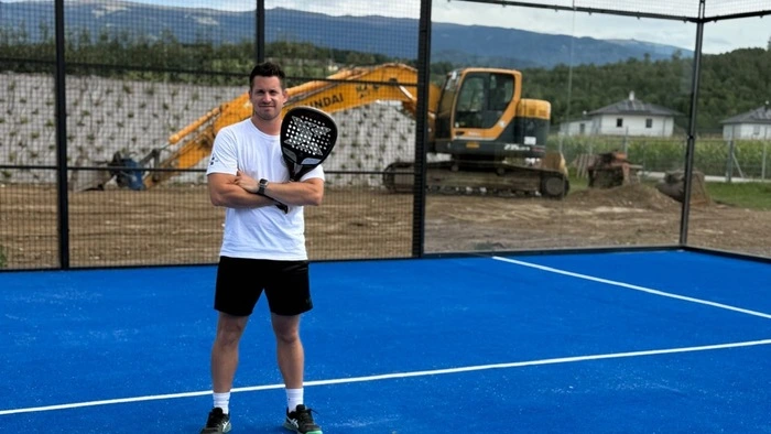 Ein Mann steht mit einem Padelschläger auf einem Padelplatz. Im Hintergrund sieht man einen Bagger.