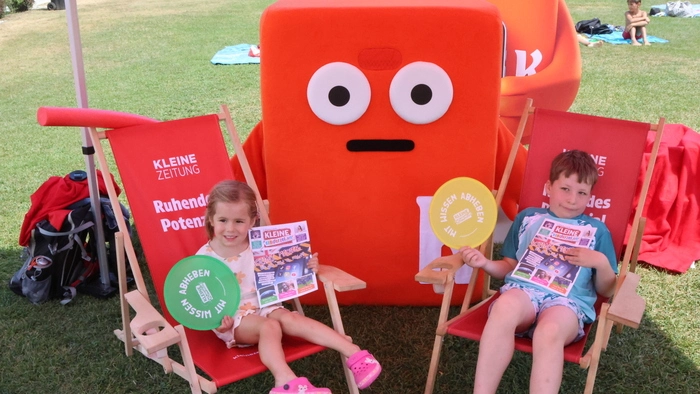 Zwei Kinder sitzen auf Strandstühlen in einem Freibad. In der Mitte steht das Kleine Zeitung-Maskottchen „Koini“.