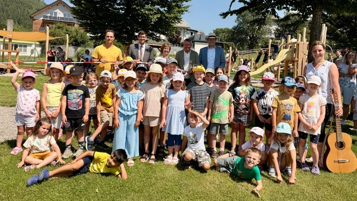 Einige Kinder sowie ein paar Erwachsene stehen vor einem Spielplatz und lächeln in die Kamera.