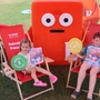 Zwei Kinder sitzen auf Strandstühlen in einem Freibad. In der Mitte steht das Kleine Zeitung-Maskottchen „Koini“.