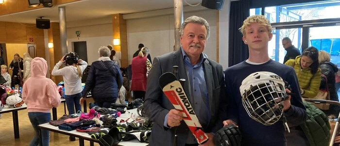 Ein älterer und ein jüngerer Mann lächeln in die Kamera. Einer hält Ski in den Händen, der andere einen Eishockey-Helm.