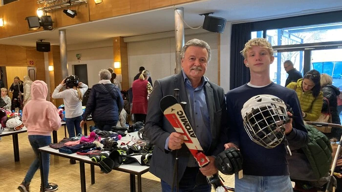 Ein älterer und ein jüngerer Mann lächeln in die Kamera. Einer hält Ski in den Händen, der andere einen Eishockey-Helm.
