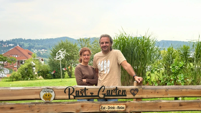 Zwei Menschen stehen hinter einem Gartenzaun mit der Aufschrift „RastGarten“
