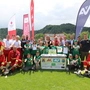 Auf einem Fußballfeld sind mehrere Kinder und Erwachsene um einen Tisch positioniert und lächeln in die Kamera