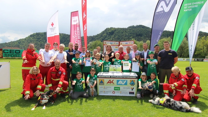 Auf einem Fußballfeld sind mehrere Kinder und Erwachsene um einen Tisch positioniert und lächeln in die Kamera