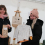Zwei Ausstellerinnen im Wirtschaftszelt am Nikolomarkt 2024. Eine hält eine Packung Müsli in der Hand, die andere eine Winterjacke für Kinder. Beide Frauen sind winterlich angezogen und lächeln in die Kamera | Lokale Aussteller sind bei der Wirtschaftsmesse anzutreffen