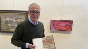 Galerist Wilfried Magnet steht in seiner Galerie und hält das Buch  „Annales Carinthiae“ aus dem Jahr 1612 in den Händen. Er hat graue Haare und trägt eine Brille. Rechts und links von ihm hängen Bilder. | Wilfried Magnet mit einem Band der „Annales Carinthiae“ von Hieronymus Megiser aus dem Jahr 1612 in den Händen. Rechts von ihm ein Bild von Martha Jungwirth