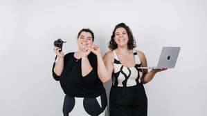 Bianca und Tamara Puschl stehen im Partnerlook gekleidet nebeneinander. Bianca hält eine Fotokamera in der Hand, Tamara einen Laptop