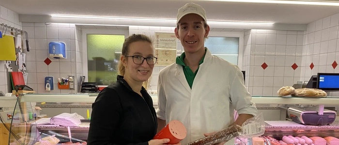 Fleischermeister Simon Čebul und seine Lebensgefährtin Yvonne Lessiak stehen vor der Fleischtheke im Geschäft. Sie ist ganz in Schwarz gekleidet, trägt eine Brille und hält eine Extrawurst in den Händen, er trägt eine weiße Kappe und einen weißen Arbeitsmantel und hält eine Salami | Fleischermeister Simon Čebul mit seiner Lebensgefährtin Yvonne Lessiak