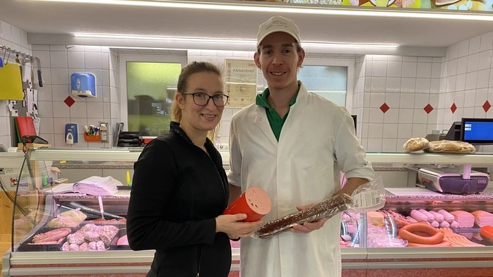 Fleischermeister Simon Čebul und seine Lebensgefährtin Yvonne Lessiak stehen vor der Fleischtheke im Geschäft. Sie ist ganz in Schwarz gekleidet, trägt eine Brille und hält eine Extrawurst in den Händen, er trägt eine weiße Kappe und einen weißen Arbeitsmantel und hält eine Salami | Fleischermeister Simon Čebul mit seiner Lebensgefährtin Yvonne Lessiak
