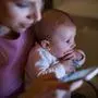 Beautiful young blond mother at home at night with her little baby son in the arms, holding smart phone, texting