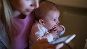 Beautiful young blond mother at home at night with her little baby son in the arms, holding smart phone, texting