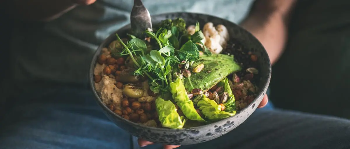 Healthy dinner or lunch. Woman in t-shirt and jeans eating vegan superbowl or Buddha bowl with hummus, vegetable, salad, beans, couscous and avocado