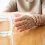 Elderly woman hands w/ tremor symptom reaching out for a glass of water on wood table. Cause of hands shaking include Parkinson's disease, stroke or brain injury. Mental health neurological disorder.