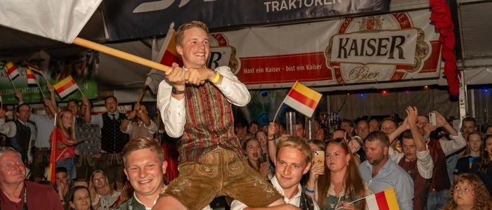 Bundessieger Gabriel Pobaschnig wird von zwei Kollegen auf den Schultern getragen und schwingt die Kärntner Fahne. Hinter ihm stehen viele Gäste, alle tragen Tracht.
