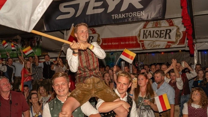 Bundessieger Gabriel Pobaschnig wird von zwei Kollegen auf den Schultern getragen und schwingt die Kärntner Fahne. Hinter ihm stehen viele Gäste, alle tragen Tracht.