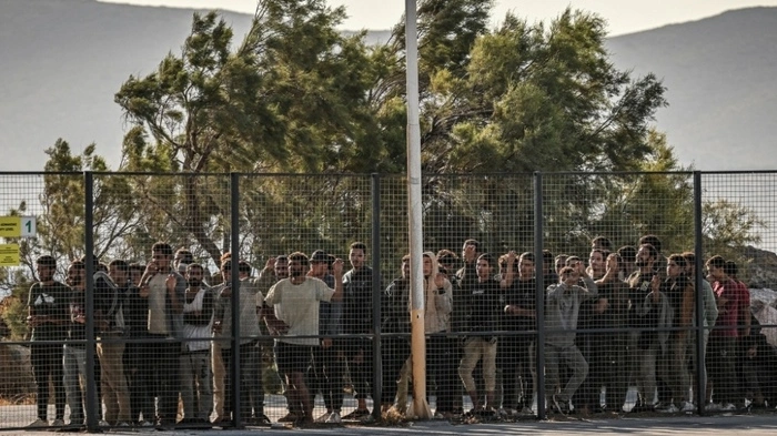 Migrants stand behinfd a fence as they are gathered by Greek coast guard officers after disembarking from a cargo ship, in the port of Lavrio, south of Athens, on July 10, 2025. The migrants were rerouted from Crete, where more than 2,000 people have arrived from Libya in recent days, sparking anger among local authorities and tourism operators on premier Kyriakos Mitsotakis' home island. (Photo by Aris MESSINIS / AFP)