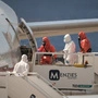 Menschen in weißen und orangefarbenen Schutzanzügen vor dem Eingang eines Flugzeugs