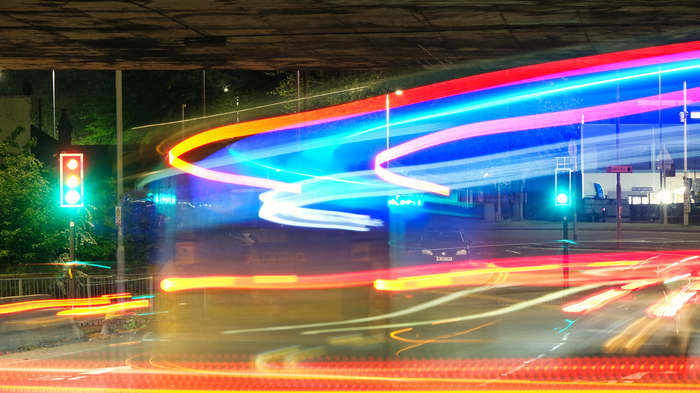 Langzeitaufnahme einer nächtlichen Straßenkreuzung unter einer Brücke. Vorbeifahrende Fahrzeuge hinterlassen rote, weiße und blaue Lichtspuren, während die Ampeln Grün zeigen.