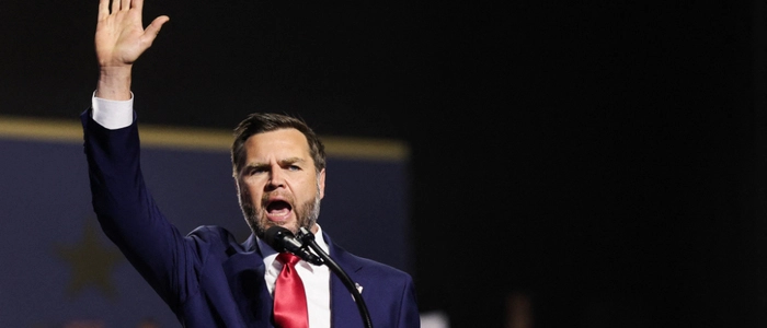 JD Vance steht an einem Rednerpult und hebt die rechte Hand.