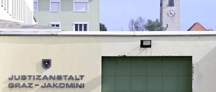 Ein Gebäude mit der Aufschrift „Justizanstalt Graz-Jakomini“ ist im Vordergrund zu sehen. Dahinter befindet sich ein grünes Haus und ein Kirchturm.