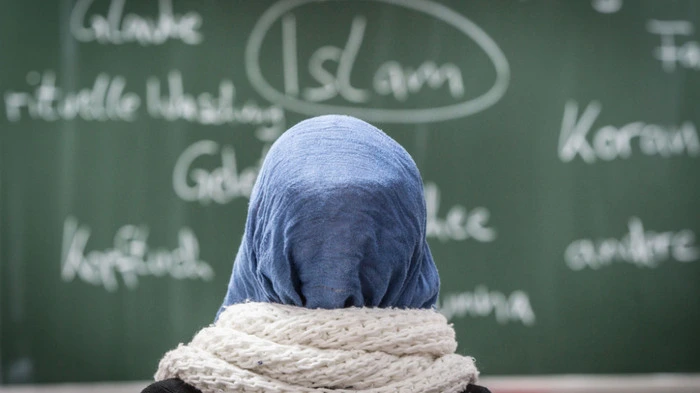 Schülerin sitzt mit Kopftuch vor einer Tafel auf der das Wort Islam eingekreist ist.