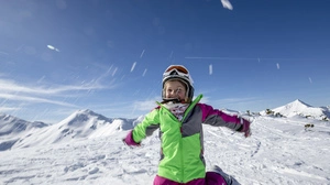 Ein Kind in einem grün-pinken Skianzug und Skihelm kniet in einer verschneiten Berglandschaft am Boden und wirft Schnee um sich