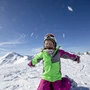 Ein Kind in einem grün-pinken Skianzug und Skihelm kniet in einer verschneiten Berglandschaft am Boden und wirft Schnee um sich
