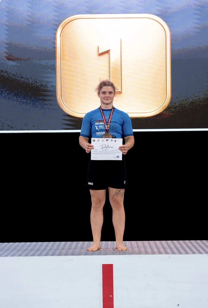 Inga Boruch steht auf einem Podium auf dem ersten Platz und hält eine Urkunde. Um ihren Hals hängt eine Medaille.