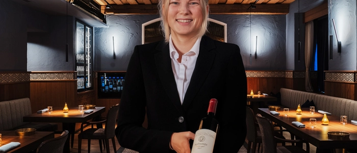 Die junge Sommelière präsentiert lächelnd eine Flasche Wein, sie trägt einen schwarzen Blazer und locker zusammengebundene blonde Haare. Im Hintergrund ein Blick in das Restaurant