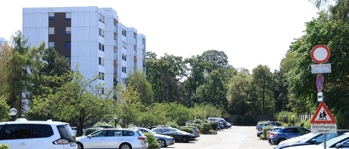 Das Foto zeigt einen langen Parkplatz, auf dem einige Autos stehen. Im Hintergrund steht ein weißes Hochhaus