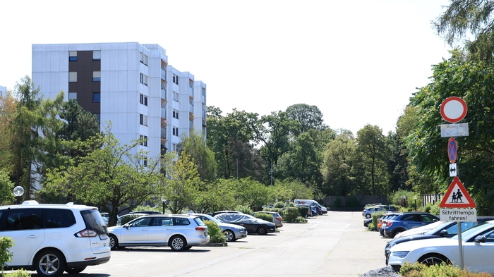 Das Foto zeigt einen langen Parkplatz, auf dem einige Autos stehen. Im Hintergrund steht ein weißes Hochhaus