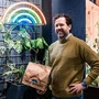 Florian Weitzer mit einer Papiertüte vor dem Regenbogen-Logo es Lokals und vielen Pflanzen an der Wand