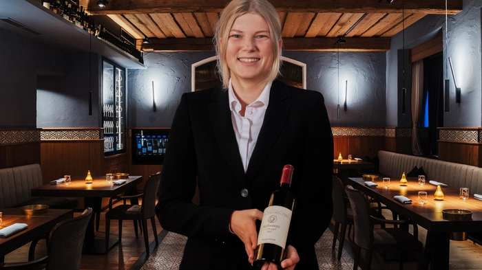 Die junge Sommelière präsentiert lächelnd eine Flasche Wein, sie trägt einen schwarzen Blazer und locker zusammengebundene blonde Haare. Im Hintergrund ein Blick in das Restaurant