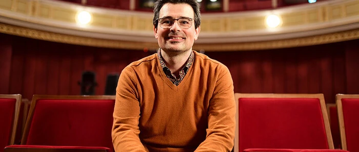 Klemens Fenllner sitz in einem orangenen Pulli und mit Brill in einem Opern- oder Kinosaal. Er lächelt freundlich in Richtung Kamera