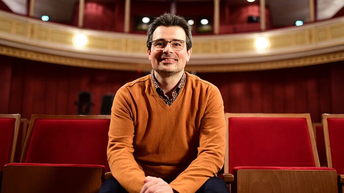 Klemens Fenllner sitz in einem orangenen Pulli und mit Brill in einem Opern- oder Kinosaal. Er lächelt freundlich in Richtung Kamera