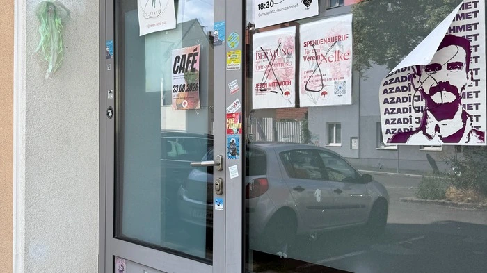 Das Bild zeigt den Eingangsbereich des Vereinslokals. Im Fenster hängen einige Poster. Davor steht die beschmierte Bank