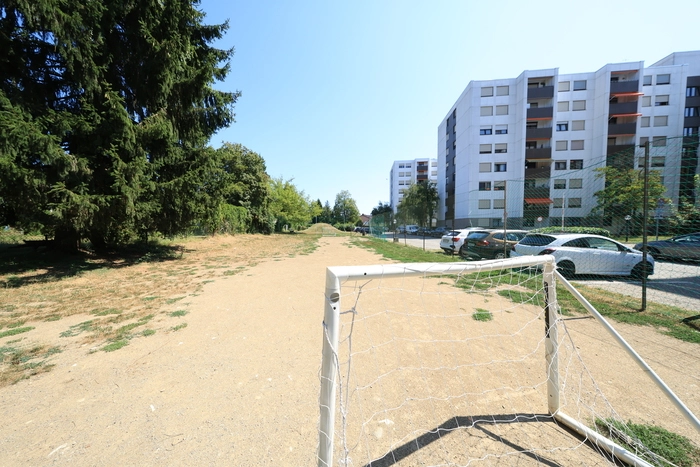 Das Bild zeigt einen Fußballplatz. Im Vordergrund steht ein Tor. Weiter hinten befindet sich der Rodelhügel. Rechts von dem Grünstreifen sind die bestehenden Parkplätze und neben ihnen die Wohnhäuser.