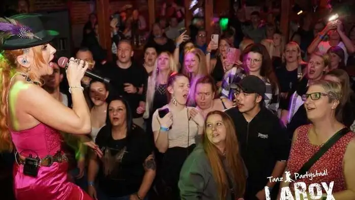 Foto aus dem Lokal: Sängerin Hannah mit Trachtenhut und rosa Kleid singt auf der Bühne, unten stehen die Fans